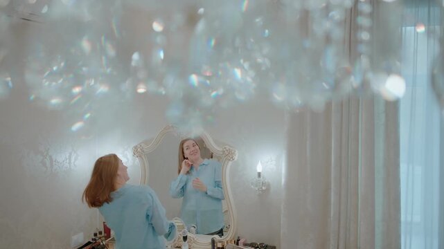 Woman in a blue robe stands before a mirror, admiring her reflection. She brushes her hair gently, smiling softly. Crystal chandelier glitters above, catching light beautifully
