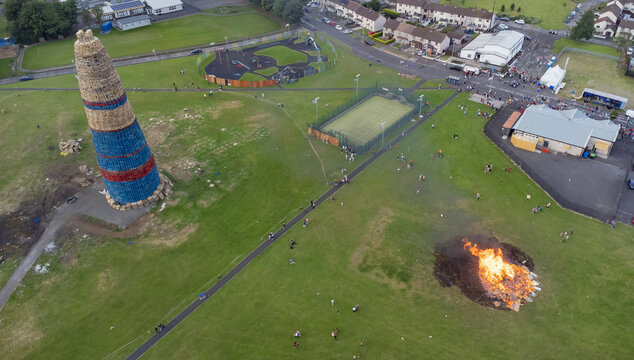 Aerial view of a large bonfire structure and a smaller burning fire in a park with a tennis court and residential houses in Larne, Northern Ireland, United Kingdom.