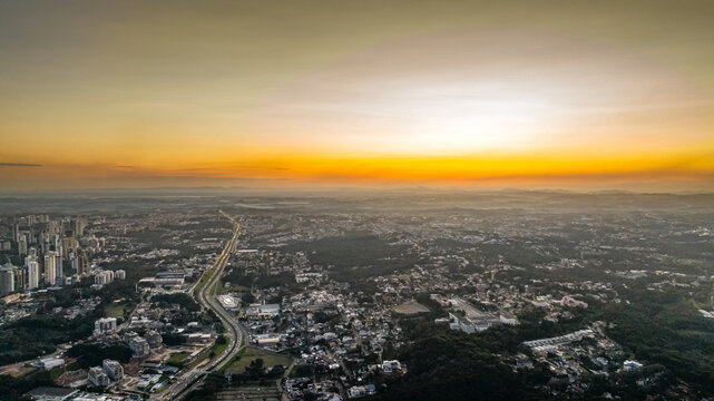 Aerial view of the city of Curitiba at sunset, featuring the urban skyline, a major highway cutting through the landscape, and dense residential areas in Curitiba, Parana, Brazil.