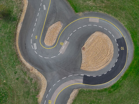 Aerial view of a vehicle testing track with asphalt curves, yellow markings, and gravel patches surrounded by green grass, United Kingdom.