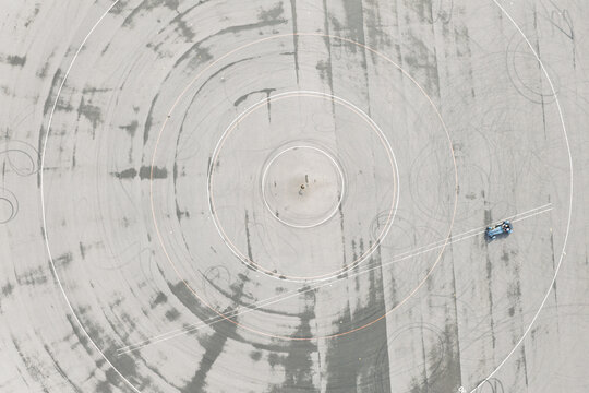 Aerial view of a blue car performing vehicle testing on a track with circular markings and tire marks on the asphalt, United Kingdom.