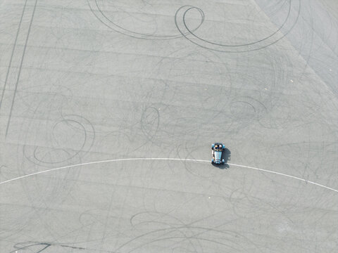 Aerial view of a sports car performing vehicle testing on a paved track marked with numerous circular tire skid marks in the United Kingdom.