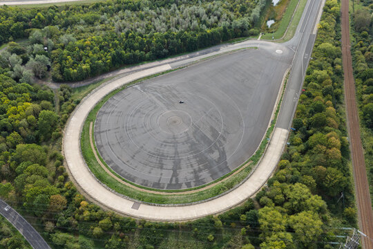 Aerial view of a vehicle testing track with a single car performing maneuvers on a large asphalt skid pad surrounded by dense green woodland and a nearby railway line in the United Kingdom.