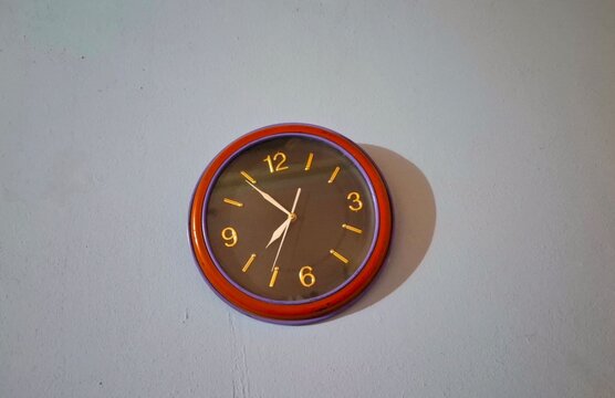 Round analog wall clock with gold numbers hanging on a plain grey wall