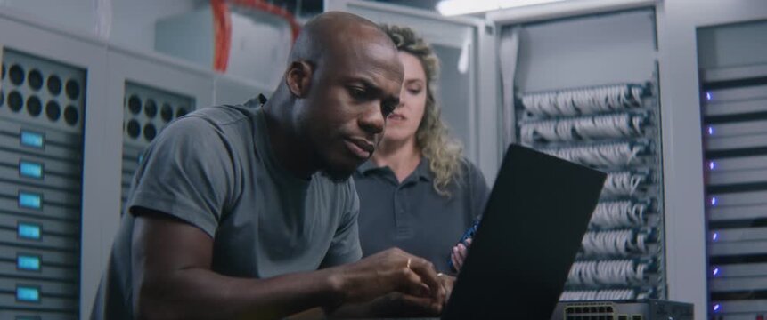 IT Specialist Analyzing Server Performance Metrics on Laptop While Discussing Diagnostic Results with Colleague. Enterprise IT Monitoring, Technical Collaboration, and Infrastructure Management.
