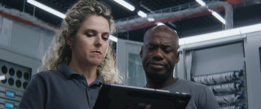 IT Specialist Presenting Network Diagnostic Logs on Digital Tablet to Male Colleague. Enterprise System Administration, Technical Collaboration, Server Configuration, IT Infrastructure Maintenance.