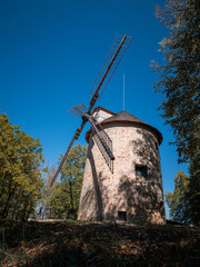 Historic 19th-century stone windmill in Holic, Slovakia, seen from the side among trees under a clear blue sky. Traditional wooden sails, rural architecture and preserved milling heritage. © Marek