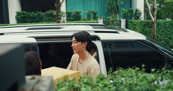 Young Asian couple unloads cardboard box from car to move into house. Family relocation home mortgage loan, Real estate vehicle transport delivery, Moving day logistics, New home arrival pack concept.