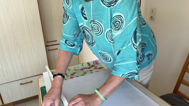 Woman in a kitchen finishes trimming the border from a large printed poster and removes the freshly cut strip of paper. The scene shows the final step of precise manual cutting. Static shot.
