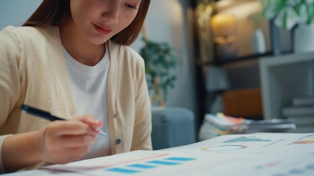 Close up young Asian woman reviews financial documents and writes budget notes at home. Personal finance planning, expense analysis, tax paperwork, household savings, banking strategy, income review.
