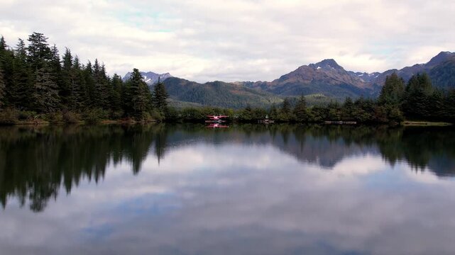 Drone flies over a mirror calm cove and past a red floatplane moored on a spruce shoreline to reveal Eyak Lake and rugged mountains near Cordova Alaska. 4K 60fps aerial footage.