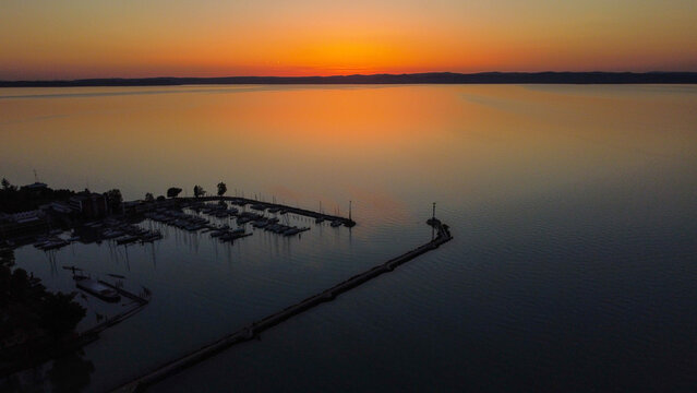 Aerial view of Lake Balaton at sunset with a marina full of sailboats, a long pier extending into the calm water, and a vibrant orange sky over the horizon in Hungary.