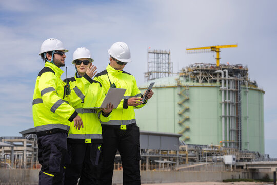 Engineers using laptop and digital tablet during site inspection near storage tank, Team of industrial inspectors in safety high-visibility jackets discussing project near gas plant facility