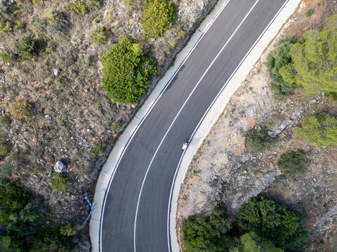 Aerial view of a cyclist riding along a winding asphalt road through the rugged landscape near Mirador del Coll de Rates de Parcent, Alicante, Valencian Community, Spain.