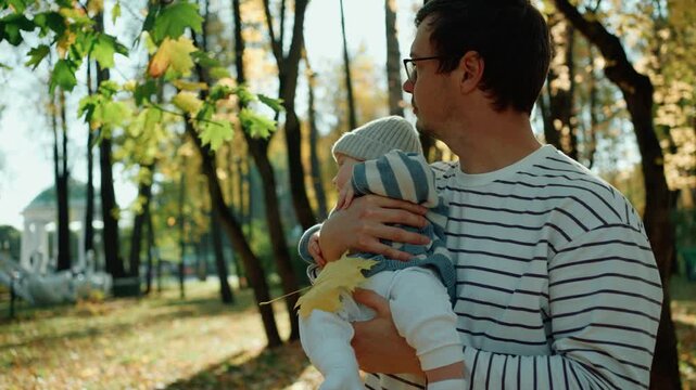 Young caring father with glasses holding his newborn son in his arms while walking through a warm, sunny autumn park, introducing the infant to the beauty of the fall season