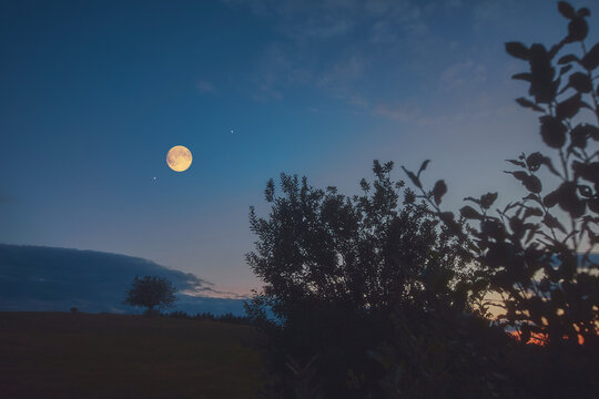 Full Moon, stars and planets above landscape silhouettes in blue hour time.