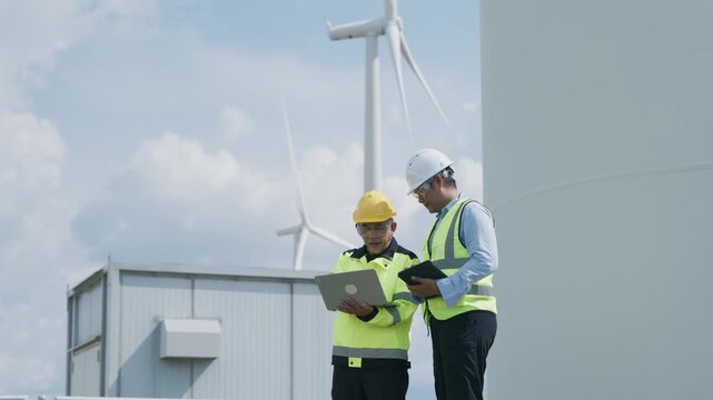 Wind Turbine Technician and Engineer Reviewing Tablet Data During Renewable Energy Farm Maintenance Inspection, Supporting Clean Power Operations and Teamwork Under Cloudy Blue Sky Conditions