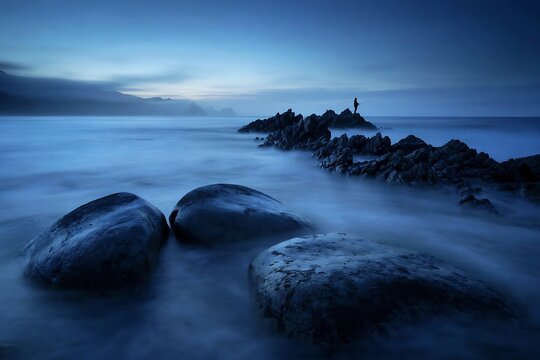 A lone fisherman stands on jagged rocks in the ocean during a serene and misty blue hour twilight