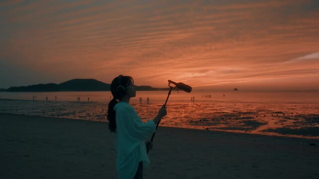 Young Woman with Headphones Holding Shotgun Microphone to Record Ambient Nature Sound at Beach Sunset