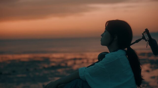 Profile View of Young Woman Sitting on Beach at Sunset with Shotgun Microphone on Shoulder