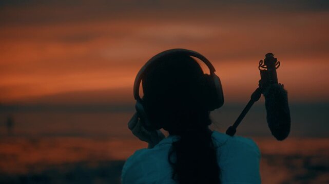 Rear View Close-Up of Woman Wearing Headphones and Holding Shotgun Microphone on Shoulder at Beach Sunset