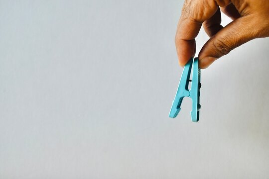 Hand holding a blue plastic clothes peg against a neutral light grey background, representing laundry accessories and pinch concepts.