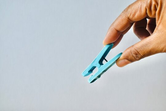 Hand holding a blue plastic clothes peg against a neutral light grey background, representing laundry accessories and pinch concepts.