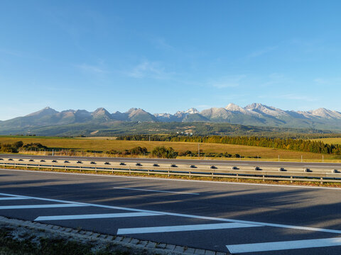 D1 motorway beneath the High Tatras in Slovakia, with snow dusted mountain peaks, autumn fields, forests and clear blue sky. Scenic road infrastructure in the transition from autumn to winter.