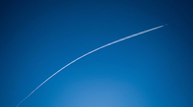 Minimalist aerial perspective of a distant jet aircraft flying at high altitude, creating a long white curved condensation trail across a solid deep azure blue sky background.