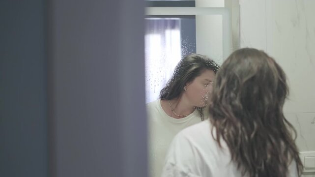 Portrait of young woman cleaning bathroom mirror with spray. Girl doing domestic chores and home maintenance. Hygiene routine, housekeeping, and lifestyle for a clean and shining interior.