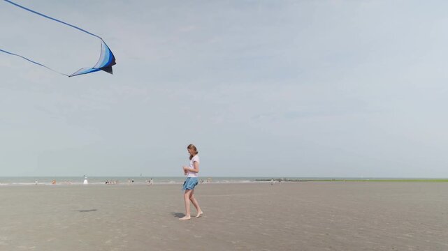 Girl flying a blue kite with long tails on a wide sandy beach walking by the sea