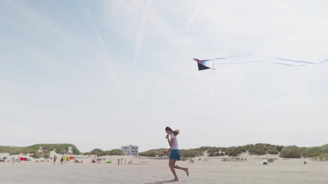 Girl running barefoot with a kite on a sandy beach slow motion sand dunes with grass in the background