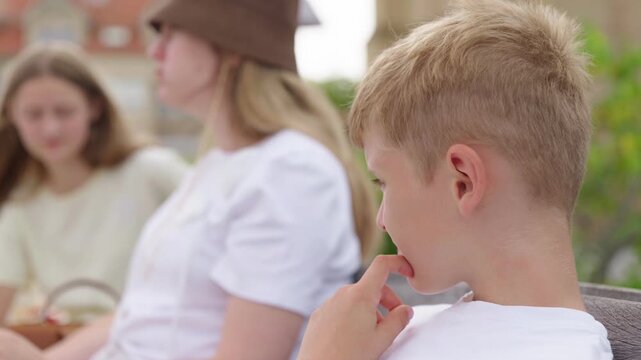 A boy is sitting on a bench next to his mother and sister slow motion a family is spending time in the park