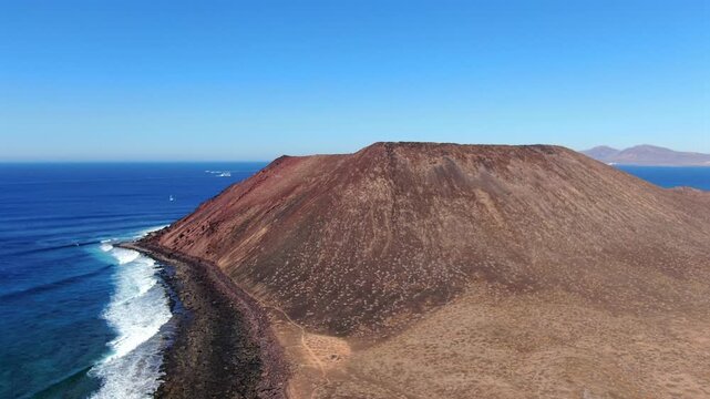 Drone flight toward the red volcanic cone of La Caldera on Isla de Lobos, with white Atlantic surf breaking along its dark lava coastline, Fuerteventura, Spain.