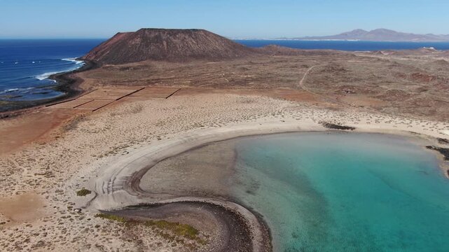 Wide aerial of the shallow turquoise lagoon of La Concha, curving between white sand and black lava, with the La Caldera volcano on the horizon, Isla de Lobos.