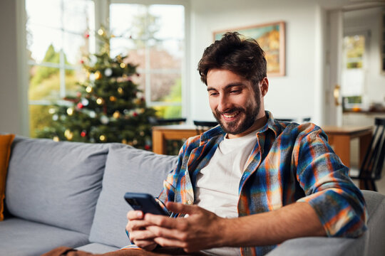 Smiling man using smartphone on sofa near christmas tree at home