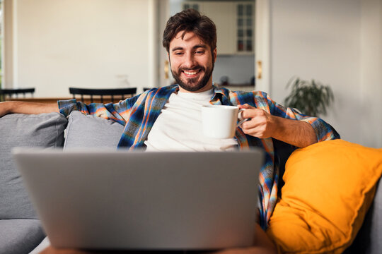 Smiling man relaxing on sofa with coffee and laptop at home