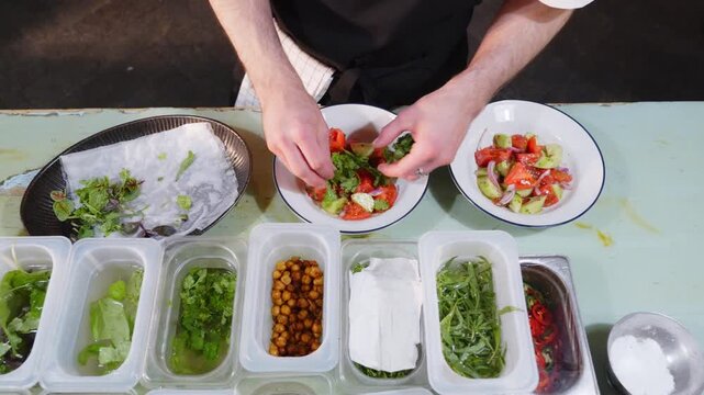 Top down view of a chef garnishing bowls of tomato, cucumber and onion salad with fresh herbs beside a row of prep containers with greens and chickpeas.