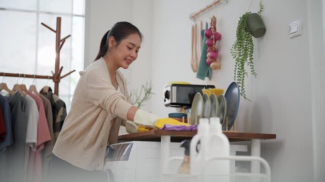 Asian Woman wiping a kitchen countertop with disinfectant spray and microfiber cloth while wearing protective gloves, promoting hygiene, sanitation, and routine home cleaning.