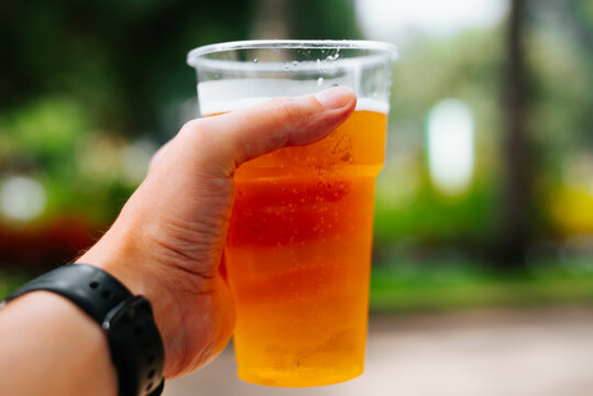 A hand wearing a black wristwatch holds a clear plastic cup filled with amber beer. Green foliage and a walkway appear out of focus in the background