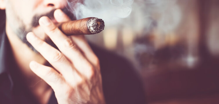 Bearded man holding a lit cigar, with smoke drifting around his face and creating a subdued atmosphere for a private tobacco ritual