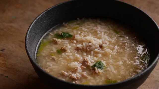 Thai rice soup with minced pork, bok choy and green onion in clear savory broth served in a dark ceramic bowl on a rustic wooden table, comforting breakfast dish