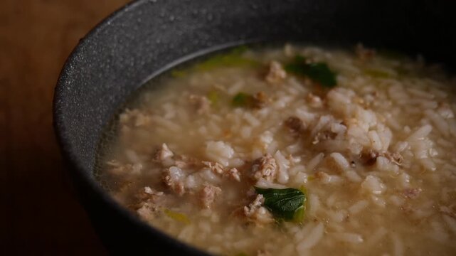 Thai rice soup with minced pork, bok choy and green onion in clear savory broth served in a dark ceramic bowl on a rustic wooden table, comforting breakfast dish