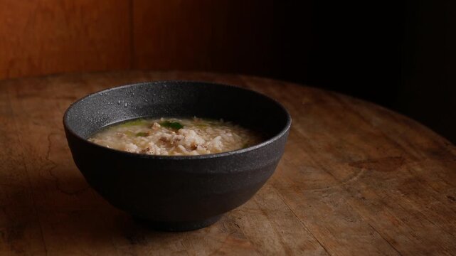 Thai rice soup with minced pork, bok choy and green onion in clear savory broth served in a dark ceramic bowl on a rustic wooden table, comforting breakfast dish