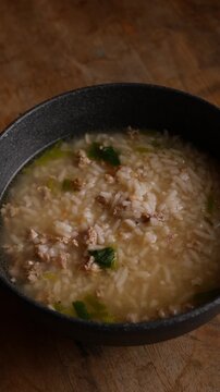 Thai rice soup with minced pork, bok choy and green onion in clear savory broth served in a dark ceramic bowl on a rustic wooden table, comforting breakfast dish