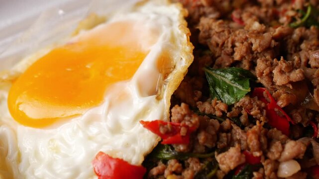 Thai basil stir-fried minced pork with crispy fried egg over steamed rice in a white foam takeaway box with plastic spoon, street food lunch on a rustic wooden table