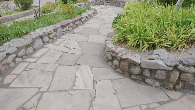Stone pathway garden landscape walkway beside rustic rock wall, gray paving through lush greenery and ornamental plant borders, peaceful outdoor setting with calm natural atmosphere for scenic travel