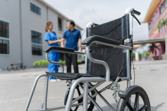 Close up of a wheelchair with blurred nurse helping senior patient to walk in background.