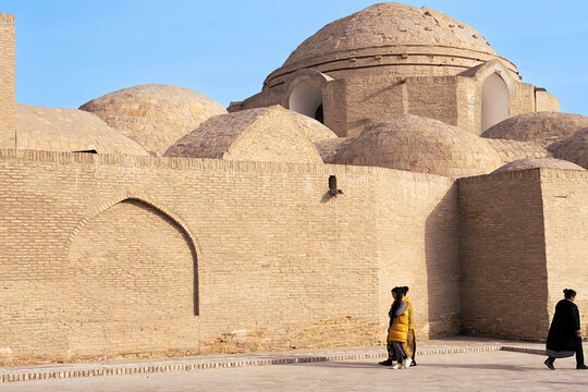 Domed brick trading dome of Toqi Zargaron in Bukhara with pedestrians walking along its massive walls. Historic Central Asian architecture under clear blue sky, ideal for travel publications.