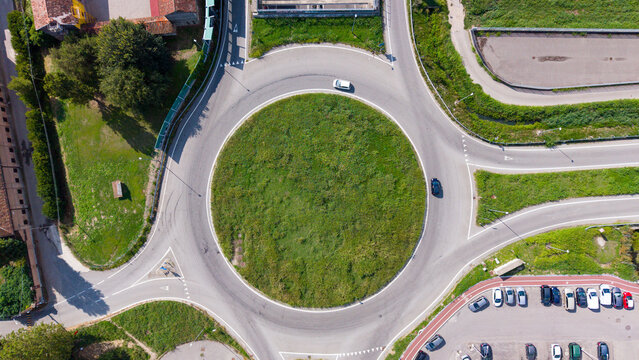 Aerial view of a circular roundabout with a green center, surrounded by roads, parked cars in a nearby lot, and surrounding greenery in Milan, Lombardy, Italy.